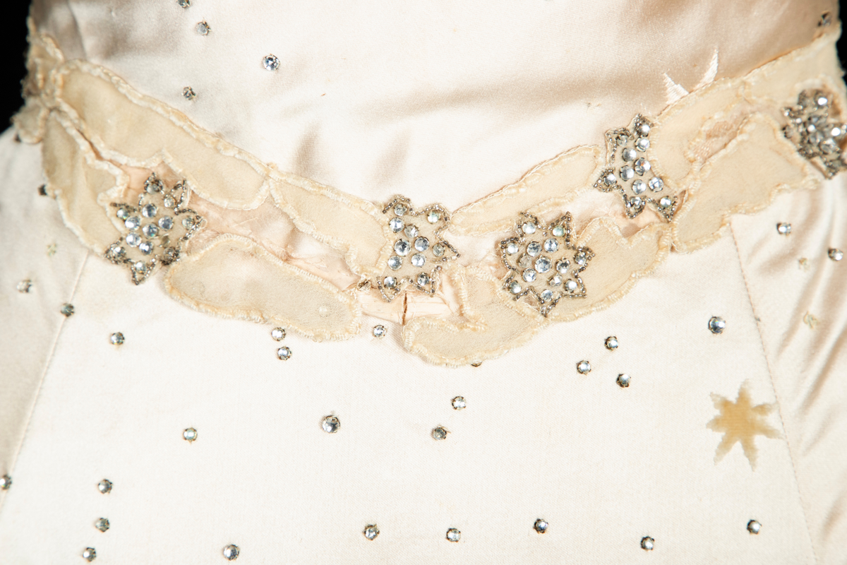 To-delt gallakjole med svært smal midje. Tettsittende, lavt ringet, kortermet livstykke og skjørt med slep. Fet, blank satengsilke, med ivevde stjerner i annen binding, deriblant stjerner av matt speilfløyel i en lys beige nyanse og skinnende blanke stjerner i en lysere nyanse. Kjolen er dekorert med små og store stjerner av begge typer, spredd utover stoffet, og i tillegg prydet med funklende "stjerner" av enkeltstående similistener/strass innfattet i blankt materiale. Kjolelivets halsringning, skulderpartier og midjeparti er også prydet med små stjerner av tettstilte similistener og noen innfelte stjerner av fløyel. Livstykkets utringning og nederkant, samt skjørtets fald og slep, er i tillegg prydet med applikerte, store og små skyer, konturert for hånd. De korte ermene og kjolelivets nederkant har kantborder av skyer sydd av chiffon, mens ringningen i tillegg er rikt dandert med bløt silketyll. Kjolelivet er avstivet med spiler, er fôret og har en innvendig lukning med snørehull midt bak. Denne er skjult av et ryggstykke som lukkes med skjulte hekter i venstre side. I tillegg har den et isydd midjebånd. Ringningen er dyp og rett skåret bak, mens fronten er asymmetrisk, med høyre skulder prydet av en stor og luftig bukett av Hortensia-blomster. Disse er utført i tekstilt materiale og delvis avstivet med metalltråd. 
Skjørtet er lett utskrådd, med vidden samlet midt bak, sluppet ut i et langt, rundet slep. Skjørtet er fôret, og har påsydd rynkekappe i nederkant av fôret. Denne er ikke faldet, men klippet i form av en tungekant.