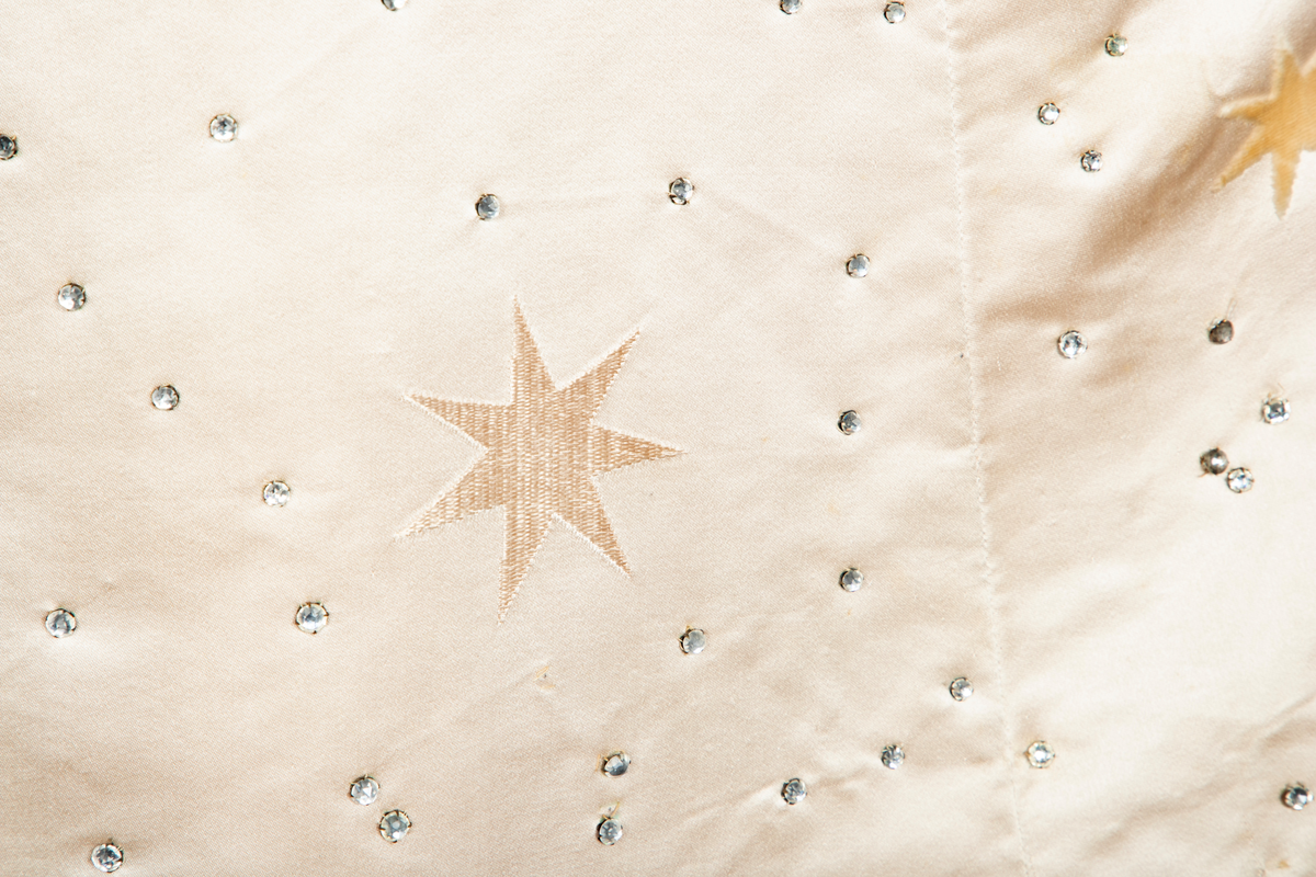 To-delt gallakjole med svært smal midje. Tettsittende, lavt ringet, kortermet livstykke og skjørt med slep. Fet, blank satengsilke, med ivevde stjerner i annen binding, deriblant stjerner av matt speilfløyel i en lys beige nyanse og skinnende blanke stjerner i en lysere nyanse. Kjolen er dekorert med små og store stjerner av begge typer, spredd utover stoffet, og i tillegg prydet med funklende "stjerner" av enkeltstående similistener/strass innfattet i blankt materiale. Kjolelivets halsringning, skulderpartier og midjeparti er også prydet med små stjerner av tettstilte similistener og noen innfelte stjerner av fløyel. Livstykkets utringning og nederkant, samt skjørtets fald og slep, er i tillegg prydet med applikerte, store og små skyer, konturert for hånd. De korte ermene og kjolelivets nederkant har kantborder av skyer sydd av chiffon, mens ringningen i tillegg er rikt dandert med bløt silketyll. Kjolelivet er avstivet med spiler, er fôret og har en innvendig lukning med snørehull midt bak. Denne er skjult av et ryggstykke som lukkes med skjulte hekter i venstre side. I tillegg har den et isydd midjebånd. Ringningen er dyp og rett skåret bak, mens fronten er asymmetrisk, med høyre skulder prydet av en stor og luftig bukett av Hortensia-blomster. Disse er utført i tekstilt materiale og delvis avstivet med metalltråd. 
Skjørtet er lett utskrådd, med vidden samlet midt bak, sluppet ut i et langt, rundet slep. Skjørtet er fôret, og har påsydd rynkekappe i nederkant av fôret. Denne er ikke faldet, men klippet i form av en tungekant.