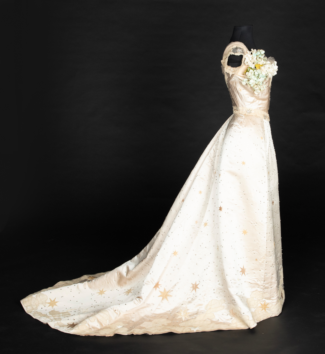 To-delt gallakjole med svært smal midje. Tettsittende, lavt ringet, kortermet livstykke og skjørt med slep. Fet, blank satengsilke, med ivevde stjerner i annen binding, deriblant stjerner av matt speilfløyel i en lys beige nyanse og skinnende blanke stjerner i en lysere nyanse. Kjolen er dekorert med små og store stjerner av begge typer, spredd utover stoffet, og i tillegg prydet med funklende "stjerner" av enkeltstående similistener/strass innfattet i blankt materiale. Kjolelivets halsringning, skulderpartier og midjeparti er også prydet med små stjerner av tettstilte similistener og noen innfelte stjerner av fløyel. Livstykkets utringning og nederkant, samt skjørtets fald og slep, er i tillegg prydet med applikerte, store og små skyer, konturert for hånd. De korte ermene og kjolelivets nederkant har kantborder av skyer sydd av chiffon, mens ringningen i tillegg er rikt dandert med bløt silketyll. Kjolelivet er avstivet med spiler, er fôret og har en innvendig lukning med snørehull midt bak. Denne er skjult av et ryggstykke som lukkes med skjulte hekter i venstre side. I tillegg har den et isydd midjebånd. Ringningen er dyp og rett skåret bak, mens fronten er asymmetrisk, med høyre skulder prydet av en stor og luftig bukett av Hortensia-blomster. Disse er utført i tekstilt materiale og delvis avstivet med metalltråd. 
Skjørtet er lett utskrådd, med vidden samlet midt bak, sluppet ut i et langt, rundet slep. Skjørtet er fôret, og har påsydd rynkekappe i nederkant av fôret. Denne er ikke faldet, men klippet i form av en tungekant.