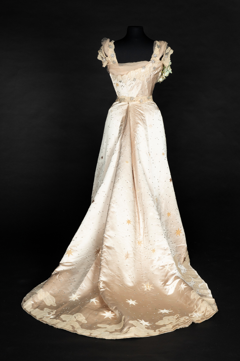 To-delt gallakjole med svært smal midje. Tettsittende, lavt ringet, kortermet livstykke og skjørt med slep. Fet, blank satengsilke, med ivevde stjerner i annen binding, deriblant stjerner av matt speilfløyel i en lys beige nyanse og skinnende blanke stjerner i en lysere nyanse. Kjolen er dekorert med små og store stjerner av begge typer, spredd utover stoffet, og i tillegg prydet med funklende "stjerner" av enkeltstående similistener/strass innfattet i blankt materiale. Kjolelivets halsringning, skulderpartier og midjeparti er også prydet med små stjerner av tettstilte similistener og noen innfelte stjerner av fløyel. Livstykkets utringning og nederkant, samt skjørtets fald og slep, er i tillegg prydet med applikerte, store og små skyer, konturert for hånd. De korte ermene og kjolelivets nederkant har kantborder av skyer sydd av chiffon, mens ringningen i tillegg er rikt dandert med bløt silketyll. Kjolelivet er avstivet med spiler, er fôret og har en innvendig lukning med snørehull midt bak. Denne er skjult av et ryggstykke som lukkes med skjulte hekter i venstre side. I tillegg har den et isydd midjebånd. Ringningen er dyp og rett skåret bak, mens fronten er asymmetrisk, med høyre skulder prydet av en stor og luftig bukett av Hortensia-blomster. Disse er utført i tekstilt materiale og delvis avstivet med metalltråd. 
Skjørtet er lett utskrådd, med vidden samlet midt bak, sluppet ut i et langt, rundet slep. Skjørtet er fôret, og har påsydd rynkekappe i nederkant av fôret. Denne er ikke faldet, men klippet i form av en tungekant.