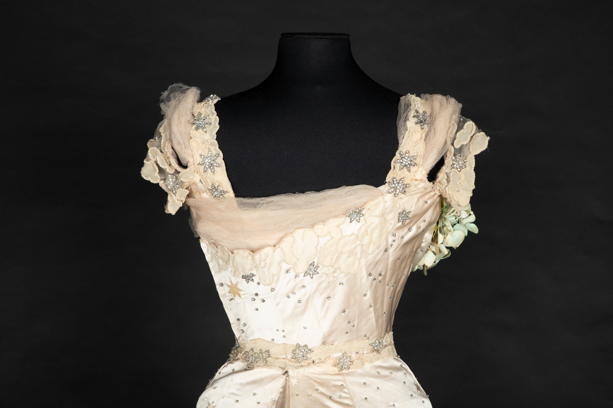 To-delt gallakjole med svært smal midje. Tettsittende, lavt ringet, kortermet livstykke og skjørt med slep. Fet, blank satengsilke, med ivevde stjerner i annen binding, deriblant stjerner av matt speilfløyel i en lys beige nyanse og skinnende blanke stjerner i en lysere nyanse. Kjolen er dekorert med små og store stjerner av begge typer, spredd utover stoffet, og i tillegg prydet med funklende "stjerner" av enkeltstående similistener/strass innfattet i blankt materiale. Kjolelivets halsringning, skulderpartier og midjeparti er også prydet med små stjerner av tettstilte similistener og noen innfelte stjerner av fløyel. Livstykkets utringning og nederkant, samt skjørtets fald og slep, er i tillegg prydet med applikerte, store og små skyer, konturert for hånd. De korte ermene og kjolelivets nederkant har kantborder av skyer sydd av chiffon, mens ringningen i tillegg er rikt dandert med bløt silketyll. Kjolelivet er avstivet med spiler, er fôret og har en innvendig lukning med snørehull midt bak. Denne er skjult av et ryggstykke som lukkes med skjulte hekter i venstre side. I tillegg har den et isydd midjebånd. Ringningen er dyp og rett skåret bak, mens fronten er asymmetrisk, med høyre skulder prydet av en stor og luftig bukett av Hortensia-blomster. Disse er utført i tekstilt materiale og delvis avstivet med metalltråd. 
Skjørtet er lett utskrådd, med vidden samlet midt bak, sluppet ut i et langt, rundet slep. Skjørtet er fôret, og har påsydd rynkekappe i nederkant av fôret. Denne er ikke faldet, men klippet i form av en tungekant.