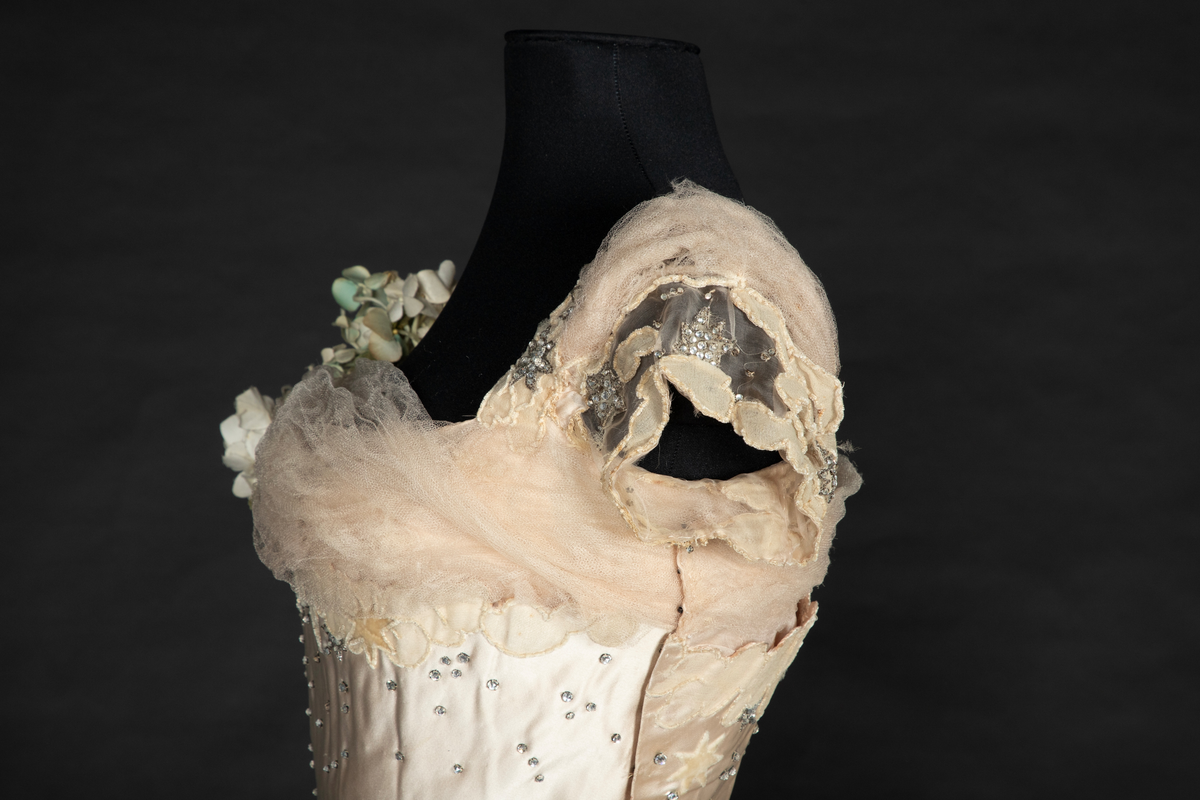 To-delt gallakjole med svært smal midje. Tettsittende, lavt ringet, kortermet livstykke og skjørt med slep. Fet, blank satengsilke, med ivevde stjerner i annen binding, deriblant stjerner av matt speilfløyel i en lys beige nyanse og skinnende blanke stjerner i en lysere nyanse. Kjolen er dekorert med små og store stjerner av begge typer, spredd utover stoffet, og i tillegg prydet med funklende "stjerner" av enkeltstående similistener/strass innfattet i blankt materiale. Kjolelivets halsringning, skulderpartier og midjeparti er også prydet med små stjerner av tettstilte similistener og noen innfelte stjerner av fløyel. Livstykkets utringning og nederkant, samt skjørtets fald og slep, er i tillegg prydet med applikerte, store og små skyer, konturert for hånd. De korte ermene og kjolelivets nederkant har kantborder av skyer sydd av chiffon, mens ringningen i tillegg er rikt dandert med bløt silketyll. Kjolelivet er avstivet med spiler, er fôret og har en innvendig lukning med snørehull midt bak. Denne er skjult av et ryggstykke som lukkes med skjulte hekter i venstre side. I tillegg har den et isydd midjebånd. Ringningen er dyp og rett skåret bak, mens fronten er asymmetrisk, med høyre skulder prydet av en stor og luftig bukett av Hortensia-blomster. Disse er utført i tekstilt materiale og delvis avstivet med metalltråd. 
Skjørtet er lett utskrådd, med vidden samlet midt bak, sluppet ut i et langt, rundet slep. Skjørtet er fôret, og har påsydd rynkekappe i nederkant av fôret. Denne er ikke faldet, men klippet i form av en tungekant.