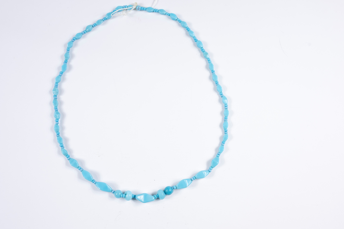Halsband med turkosa plastpärlor i större och mindre runda respektive avlånga och kantiga former. De största pärlorna mitt fram. Pärlorna är trädda på ett tandtrådsliknande material, troligtvis har den ursprungliga tråden gått av och "tandtråden" har fått ersätta denna. Halsbandet saknar låsanordning.