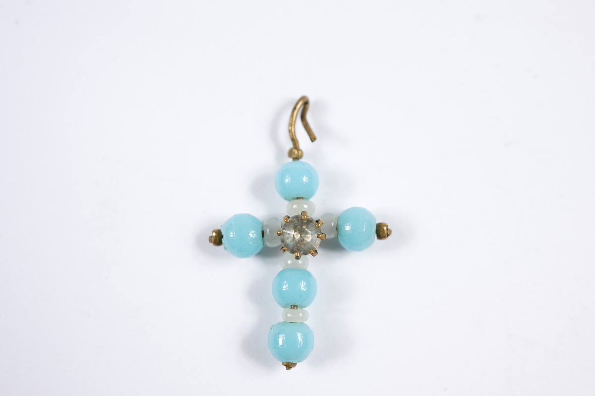 Hängsmycke i form av ett kors. Turkosa plastpärlor och vita glaspärlor trädda på metalltråd som formats till ett kors. Där korsarmarna möts sitter en plast- eller glaspärla som slipats för att efterlikna en ädelsten i en infattning.