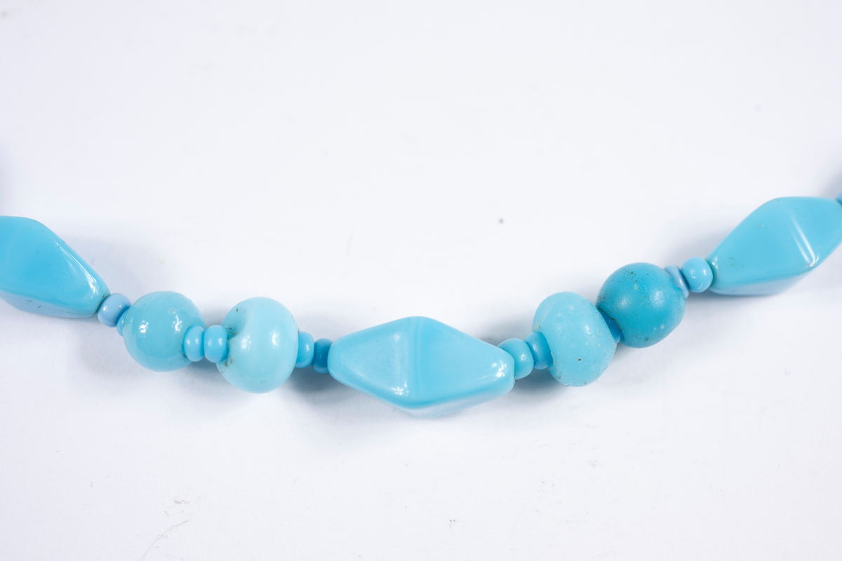 Halsband med turkosa plastpärlor i större och mindre runda respektive avlånga och kantiga former. De största pärlorna mitt fram. Pärlorna är trädda på ett tandtrådsliknande material, troligtvis har den ursprungliga tråden gått av och "tandtråden" har fått ersätta denna. Halsbandet saknar låsanordning.