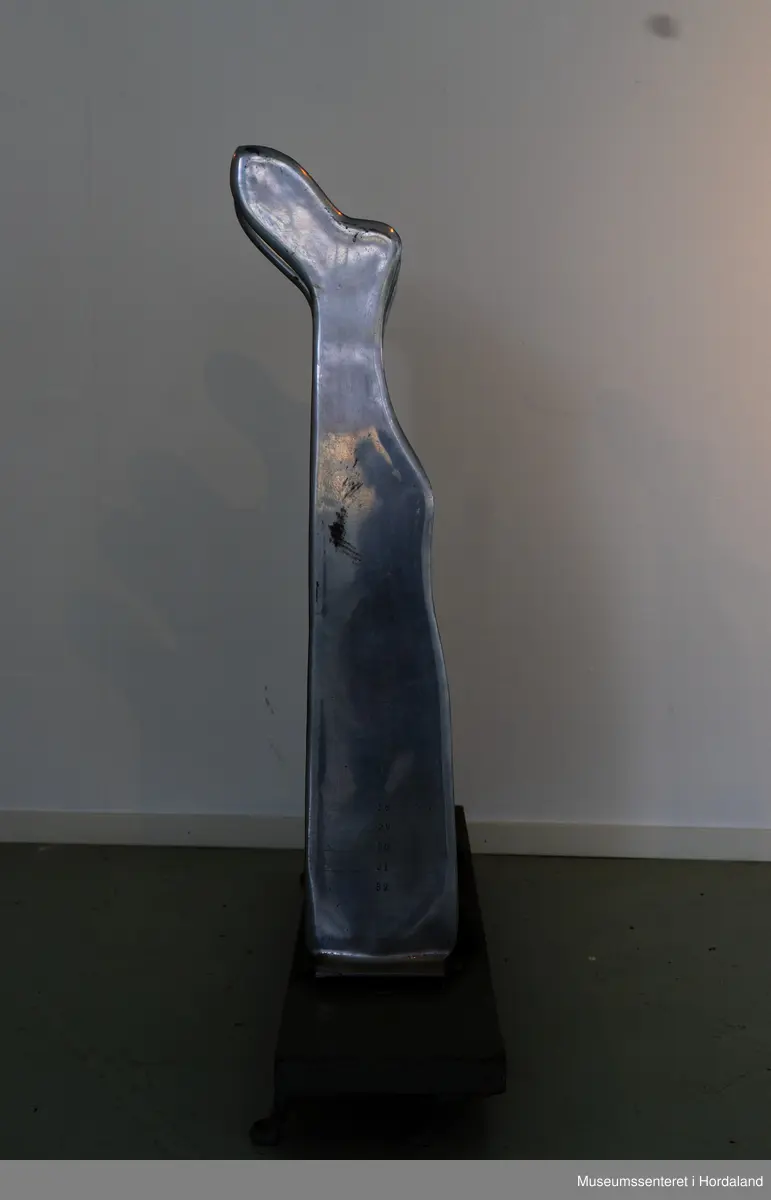 Fire strømpeformer i metall skrudd fast til rektangulær treplanke på fire metallføtter og to runde, svarte brytere av bakelitt. Strømpeformene er flate og formet som en fot. Treplate og metallføtter er gråmalte.