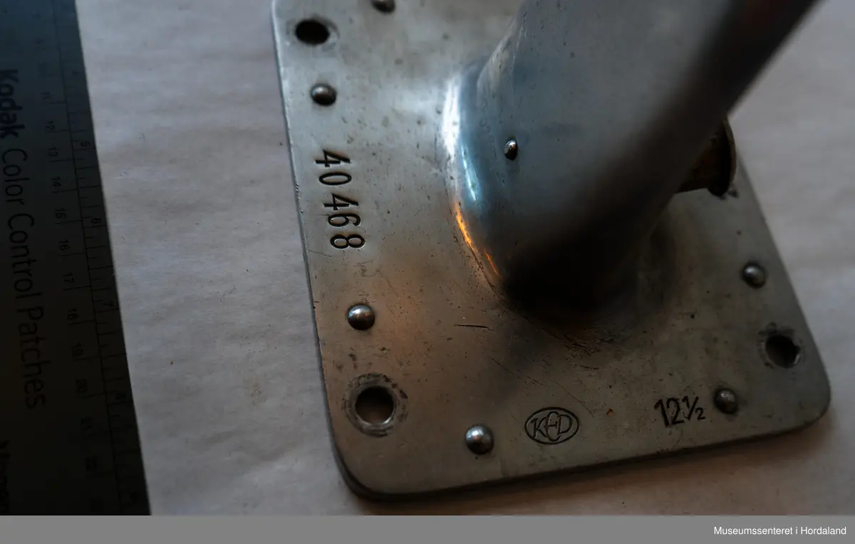 Sokkeform i metall. Den er flat og formet som en fot. Formen står opp fra en flat, rektangulær plate med hull i hvert hjørne.