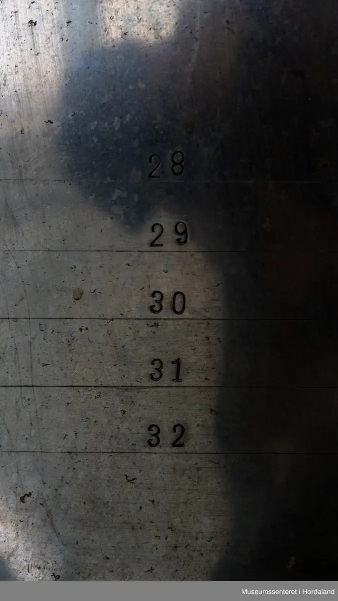 Fire strømpeformer i metall skrudd fast til rektangulær treplanke på fire metallføtter og to runde, svarte brytere av bakelitt. Strømpeformene er flate og formet som en fot. Treplate og metallføtter er gråmalte.