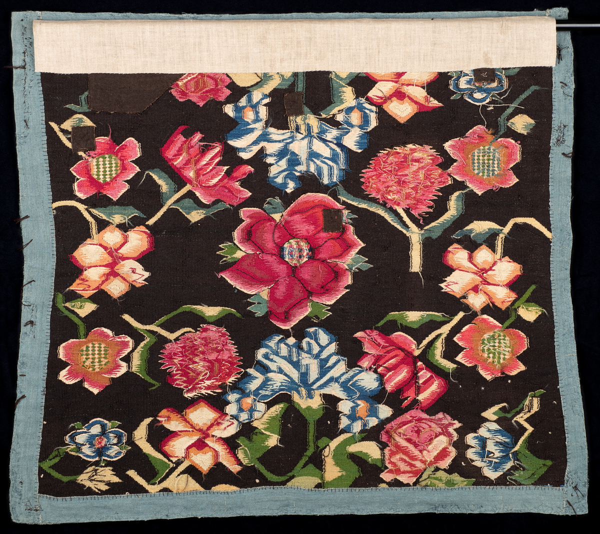Putetrekk av billedvev. Renning av lin og innslag av ull (bomull?). Røde roser, rosa markblomster og blå liljer med grønne og hvite blader på mørk bunn.