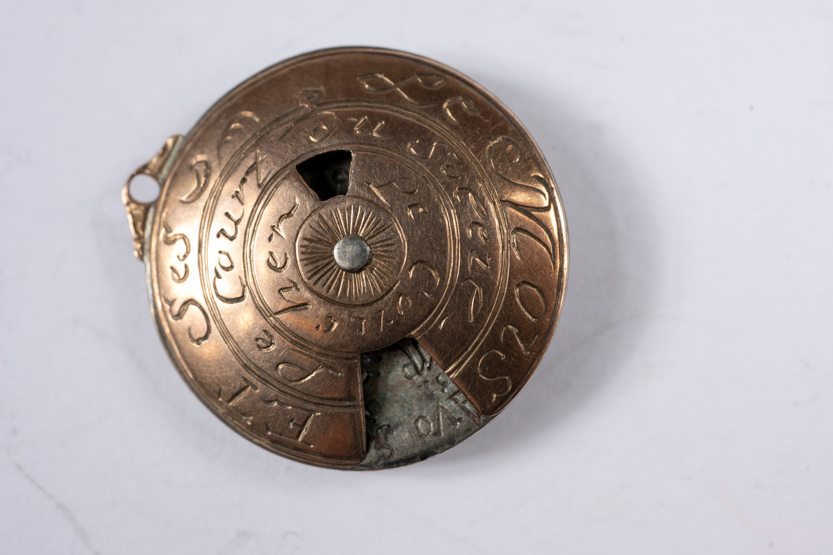 Vit och rödaktig metall, möjligen järn och koppar. Graverad på franska med månader, dagar, datum och zodiaksymboler. Gulddoublé kedja, länk och stav. 700 mm lång.