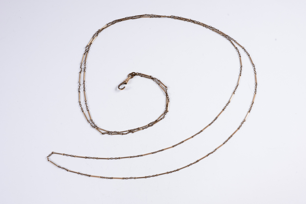 Vit och rödaktig metall, möjligen järn och koppar. Graverad på franska med månader, dagar, datum och zodiaksymboler. Gulddoublé kedja, länk och stav. 700 mm lång.