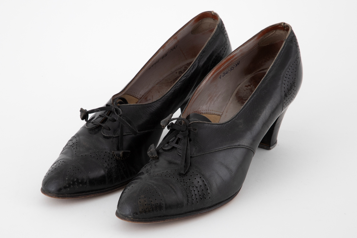 Snøresko i svart skinn med lærhæl og lærsåle. Skoene har lav snøring med 3 x 3 hull, tilspisset tå og halvhøy, lett svunget hæl. Perforert dekor. Lisser av bånd med skinnrosetter som endestykker. Fôr av lyst grått skinn, i tåen av stoff.