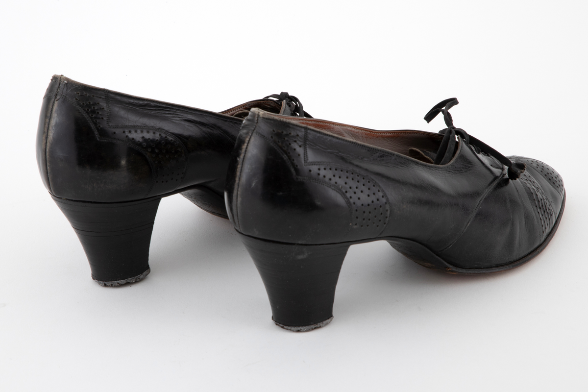 Snøresko i svart skinn med lærhæl og lærsåle. Skoene har lav snøring med 3 x 3 hull, tilspisset tå og halvhøy, lett svunget hæl. Perforert dekor. Lisser av bånd med skinnrosetter som endestykker. Fôr av lyst grått skinn, i tåen av stoff.