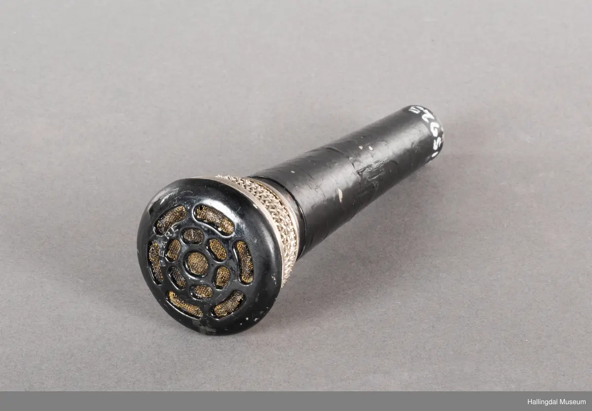 Mikrofon av merket Pearl. Svart kjegleformet håndtak med kabelkontakt i bunnen. Håndtaket kan se svartlakkert ut, mulig av bruker. Mikrofongrillen er av både metall og plast. 