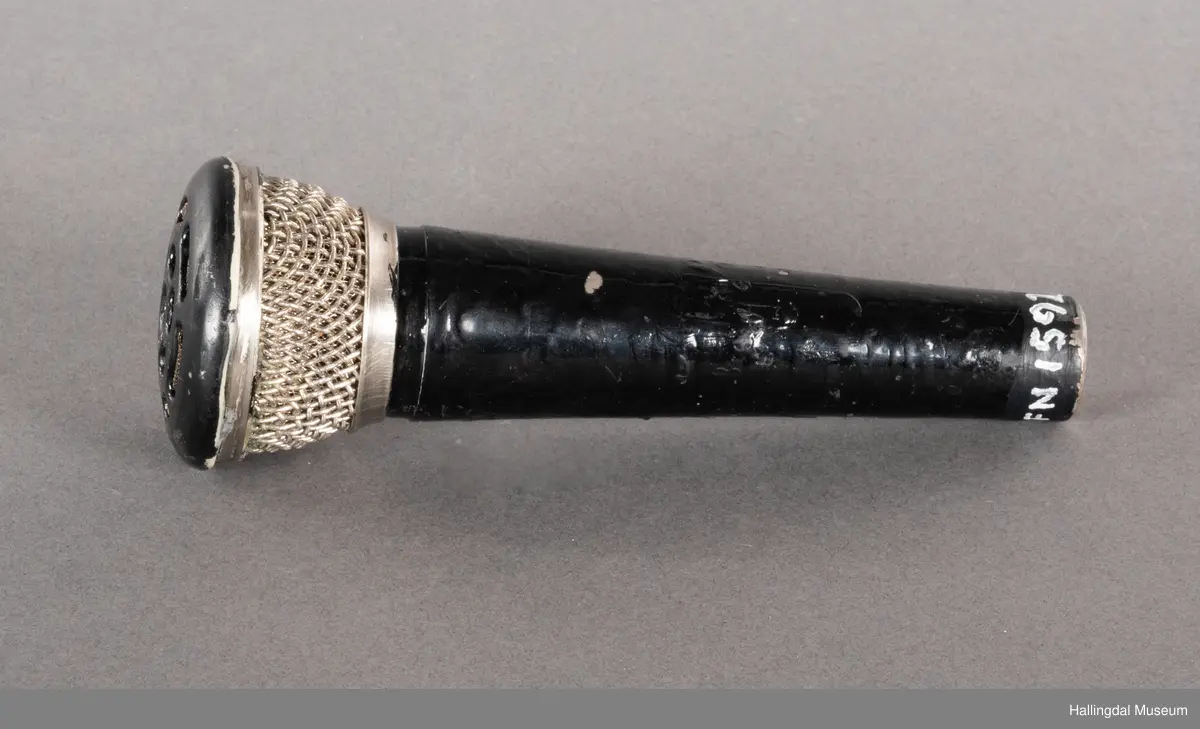 Mikrofon av merket Pearl. Svart kjegleformet håndtak med kabelkontakt i bunnen. Håndtaket kan se svartlakkert ut, mulig av bruker. Mikrofongrillen er av både metall og plast. 