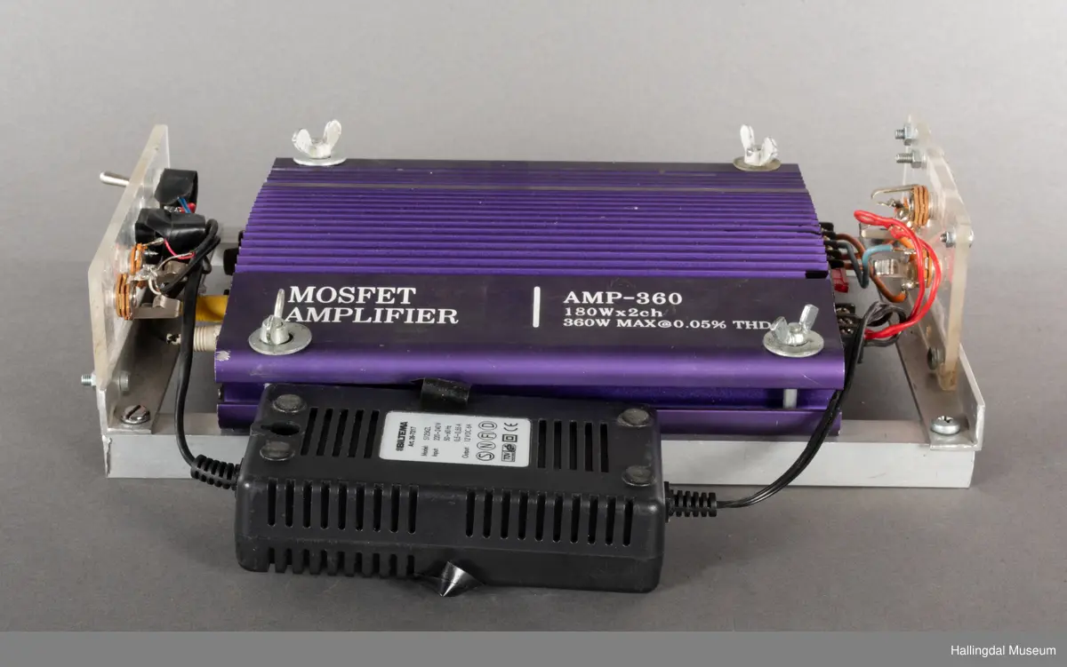 Hjemmelaget transportabelt lydanlegg. Rødbrun skinnkoffert med messingbeslag. Lysebrunt skinn innvendig fôr. Inneholder en ombygd "Mosfet" amplifier i lilla farge, med tilhørende grå strømkabel. På sidene er det plexiglass. Høyre side plexiglass bestående av strøminntak kontakt og en av og på bryter og to stk. merket mikserinngang til jack. På venstre side plexiglass inntak med merke: 12V - DG og to høytalerinnganger. Kabler til høytaler og mikrofon mangler.  