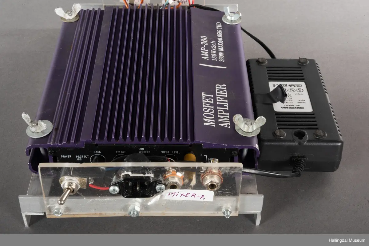 Hjemmelaget transportabelt lydanlegg. Rødbrun skinnkoffert med messingbeslag. Lysebrunt skinn innvendig fôr. Inneholder en ombygd "Mosfet" amplifier i lilla farge, med tilhørende grå strømkabel. På sidene er det plexiglass. Høyre side plexiglass bestående av strøminntak kontakt og en av og på bryter og to stk. merket mikserinngang til jack. På venstre side plexiglass inntak med merke: 12V - DG og to høytalerinnganger. Kabler til høytaler og mikrofon mangler.  
