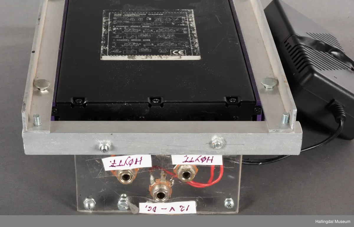 Hjemmelaget transportabelt lydanlegg. Rødbrun skinnkoffert med messingbeslag. Lysebrunt skinn innvendig fôr. Inneholder en ombygd "Mosfet" amplifier i lilla farge, med tilhørende grå strømkabel. På sidene er det plexiglass. Høyre side plexiglass bestående av strøminntak kontakt og en av og på bryter og to stk. merket mikserinngang til jack. På venstre side plexiglass inntak med merke: 12V - DG og to høytalerinnganger. Kabler til høytaler og mikrofon mangler.  
