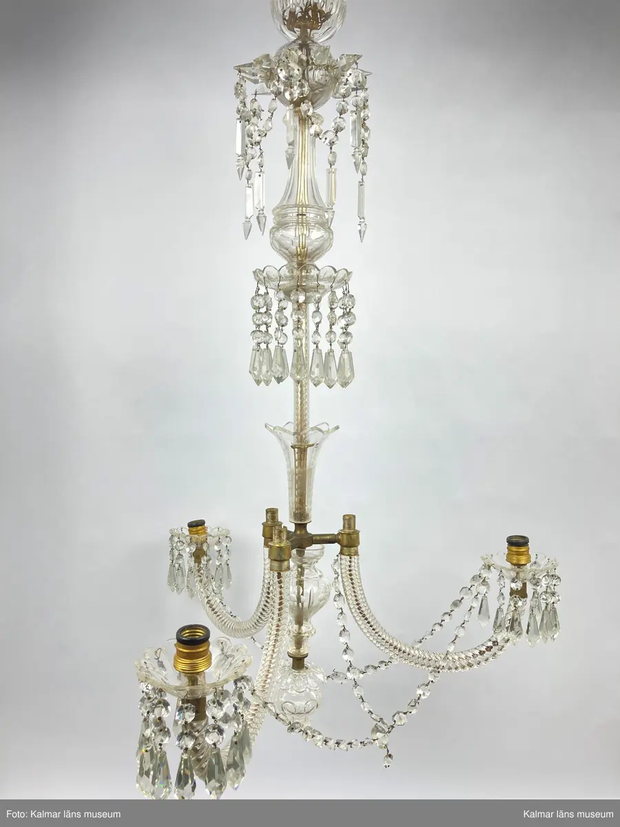 KLM 46969:1. Ljuskrona. Venetiansk stil. Stomme av mässing, övriga detaljer av slipat glas i olika former. Elektrifierad. På metallstommen en stämpel: EP. Till ljuskronan hör diverse lösa delar, se KLM 46969:4.