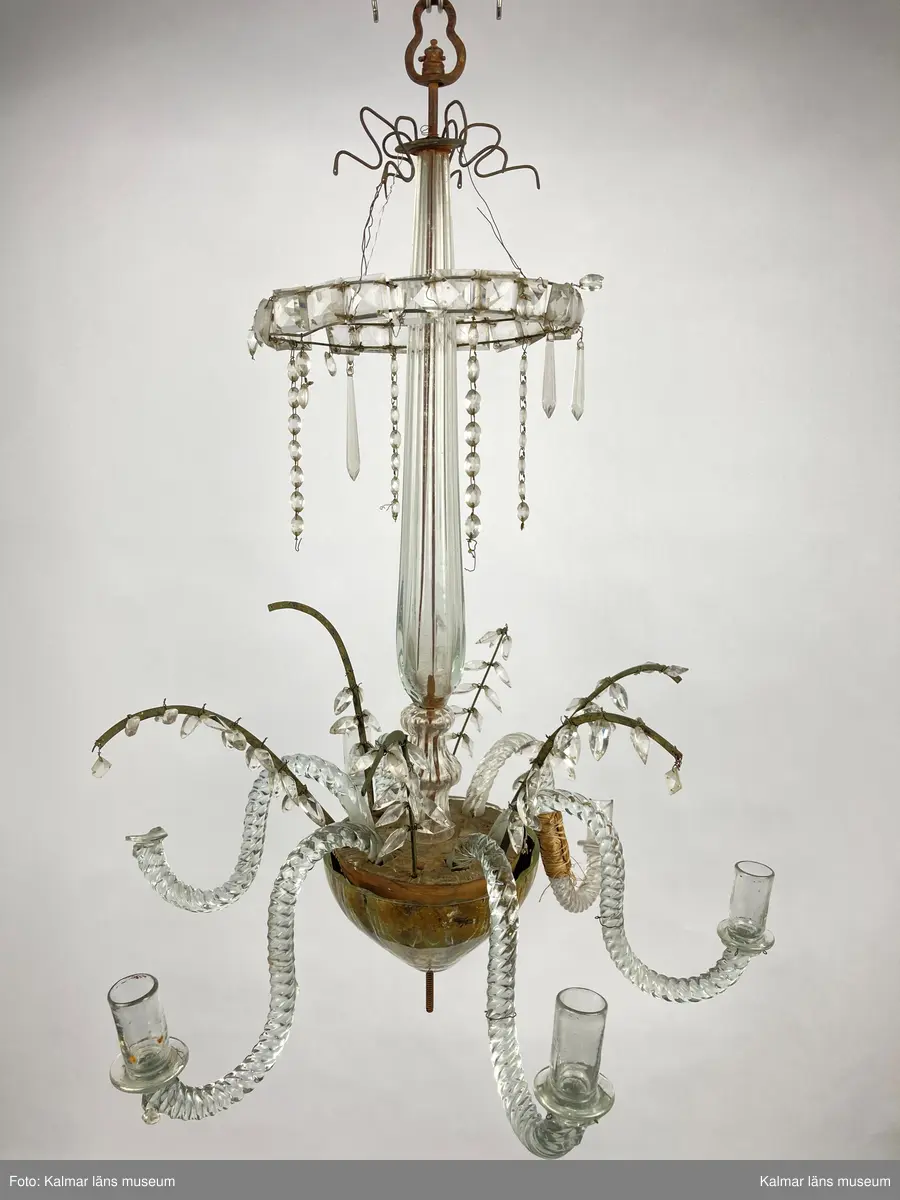 KLM 46968. Ljuskrona. Stomme av metall och trä med löstagbara ljusarmar av spiralvridet glas. Även löstagbara metallbågar med prismor. Nedre glaspartiet belagt med guldfärg på insidan. Prismor av slipat glas, flertalet lösa och osäkert hur de har suttit. Ena ljusarmen har en äldre lagning.