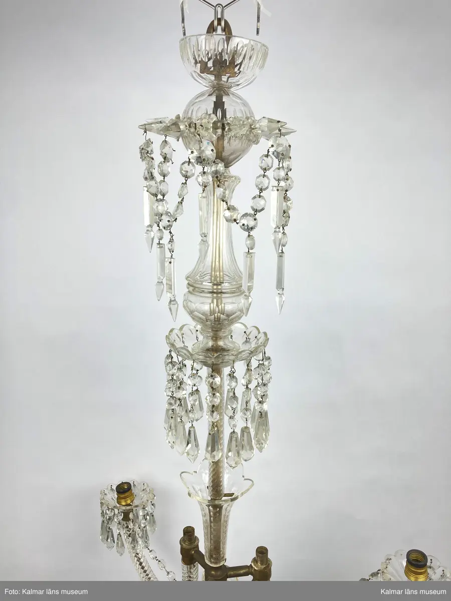 KLM 46969:1. Ljuskrona. Venetiansk stil. Stomme av mässing, övriga detaljer av slipat glas i olika former. Elektrifierad. På metallstommen en stämpel: EP. Till ljuskronan hör diverse lösa delar, se KLM 46969:4.