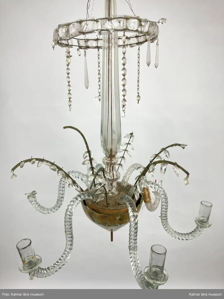 KLM 46968. Ljuskrona. Stomme av metall och trä med löstagbara ljusarmar av spiralvridet glas. Även löstagbara metallbågar med prismor. Nedre glaspartiet belagt med guldfärg på insidan. Prismor av slipat glas, flertalet lösa och osäkert hur de har suttit. Ena ljusarmen har en äldre lagning.