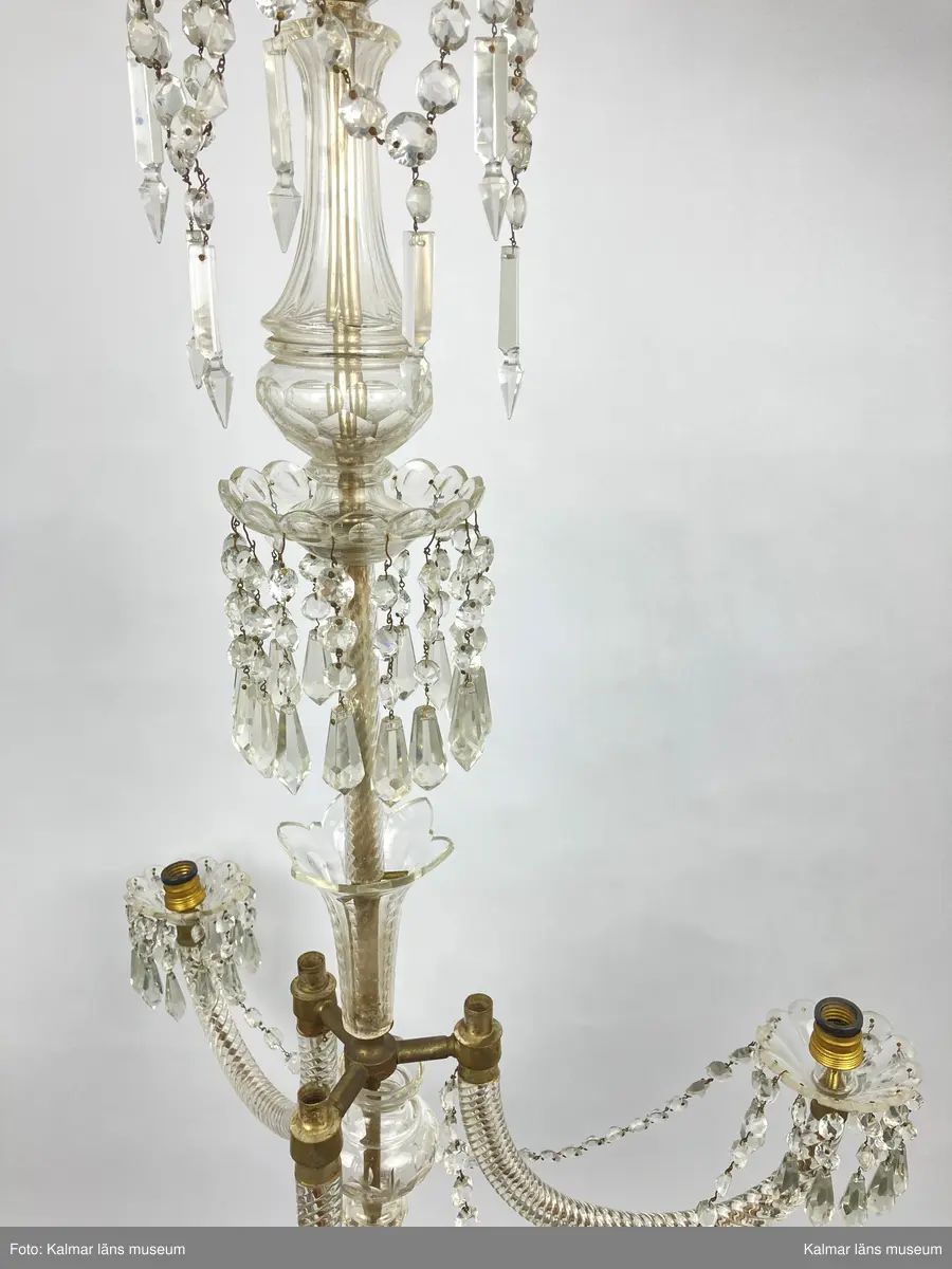 KLM 46969:1. Ljuskrona. Venetiansk stil. Stomme av mässing, övriga detaljer av slipat glas i olika former. Elektrifierad. På metallstommen en stämpel: EP. Till ljuskronan hör diverse lösa delar, se KLM 46969:4.