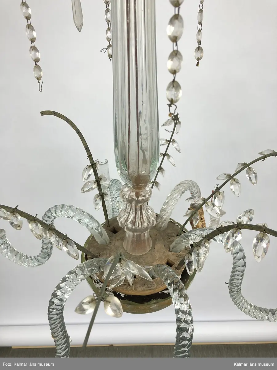 KLM 46968. Ljuskrona. Stomme av metall och trä med löstagbara ljusarmar av spiralvridet glas. Även löstagbara metallbågar med prismor. Nedre glaspartiet belagt med guldfärg på insidan. Prismor av slipat glas, flertalet lösa och osäkert hur de har suttit. Ena ljusarmen har en äldre lagning.