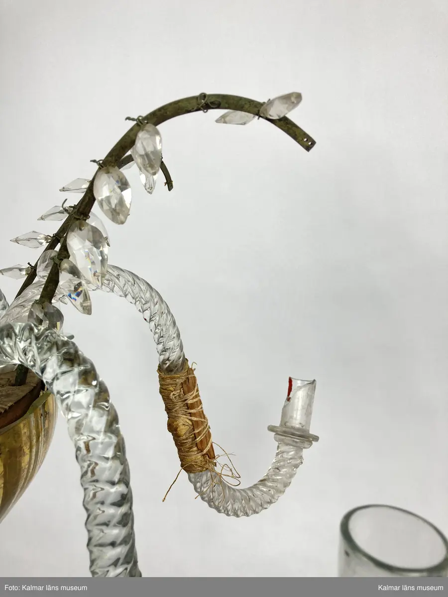 KLM 46968. Ljuskrona. Stomme av metall och trä med löstagbara ljusarmar av spiralvridet glas. Även löstagbara metallbågar med prismor. Nedre glaspartiet belagt med guldfärg på insidan. Prismor av slipat glas, flertalet lösa och osäkert hur de har suttit. Ena ljusarmen har en äldre lagning.