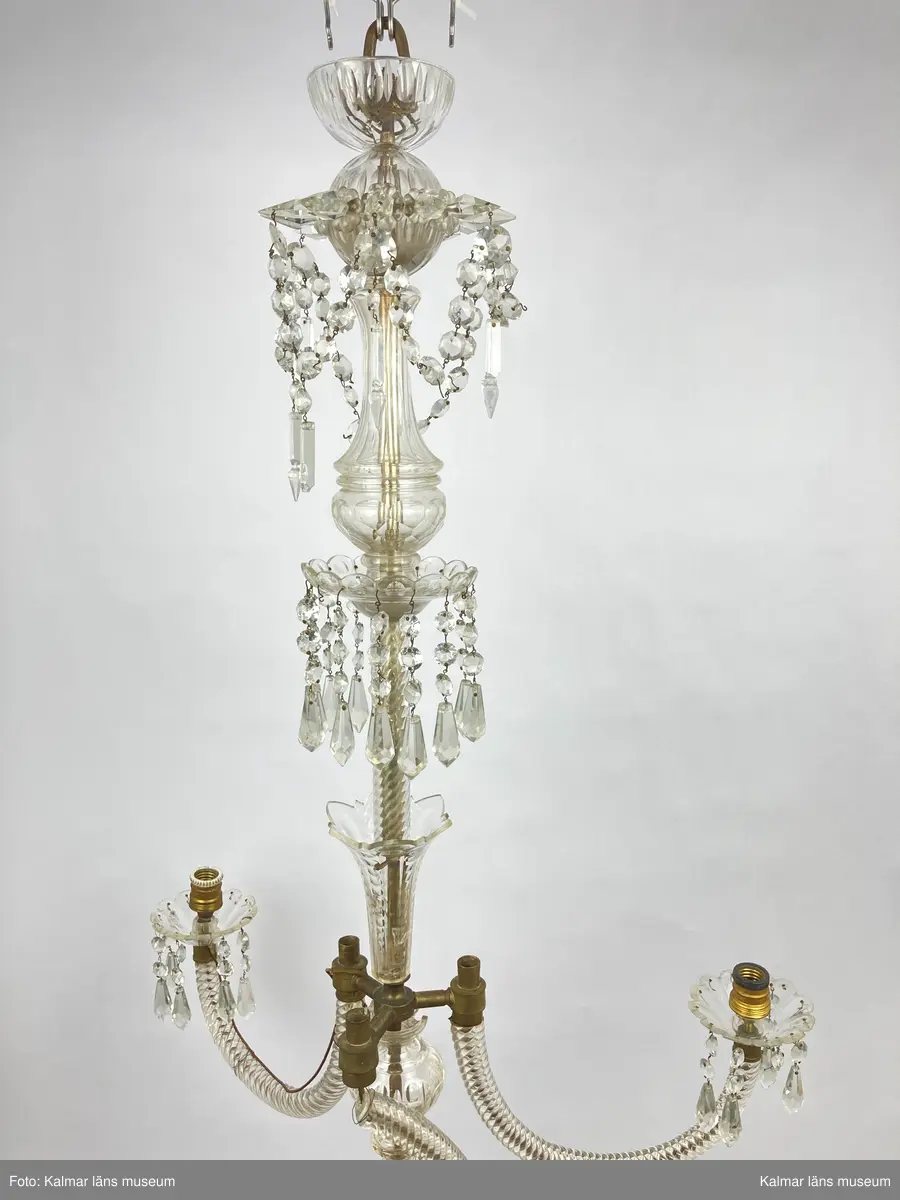 KLM 46969:3. Ljuskrona. Venetiansk stil. Stomme av mässing, övriga detaljer av slipat glas i olika former. Elektrifierad. Till ljuskronan hör diverse lösa delar, se KLM 46969:4.