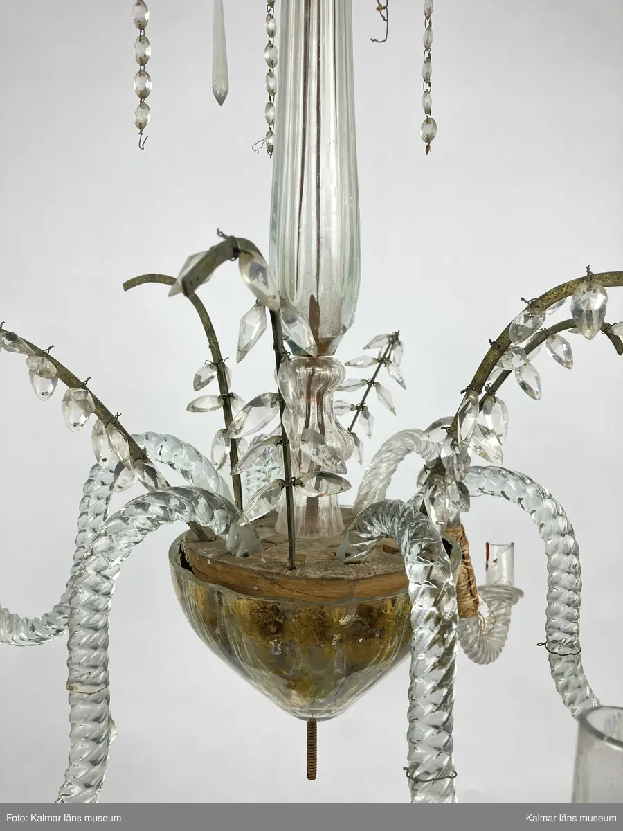 KLM 46968. Ljuskrona. Stomme av metall och trä med löstagbara ljusarmar av spiralvridet glas. Även löstagbara metallbågar med prismor. Nedre glaspartiet belagt med guldfärg på insidan. Prismor av slipat glas, flertalet lösa och osäkert hur de har suttit. Ena ljusarmen har en äldre lagning.