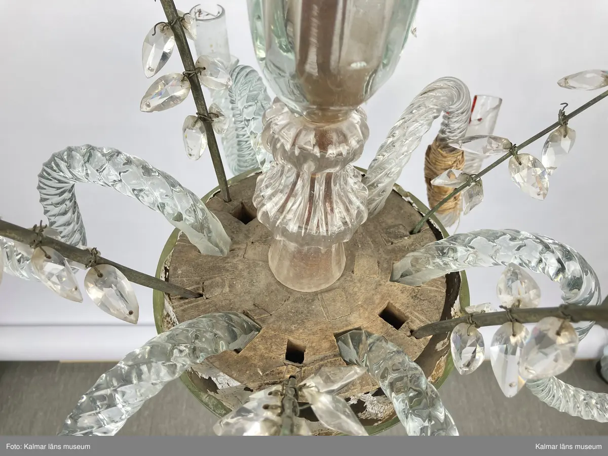 KLM 46968. Ljuskrona. Stomme av metall och trä med löstagbara ljusarmar av spiralvridet glas. Även löstagbara metallbågar med prismor. Nedre glaspartiet belagt med guldfärg på insidan. Prismor av slipat glas, flertalet lösa och osäkert hur de har suttit. Ena ljusarmen har en äldre lagning.