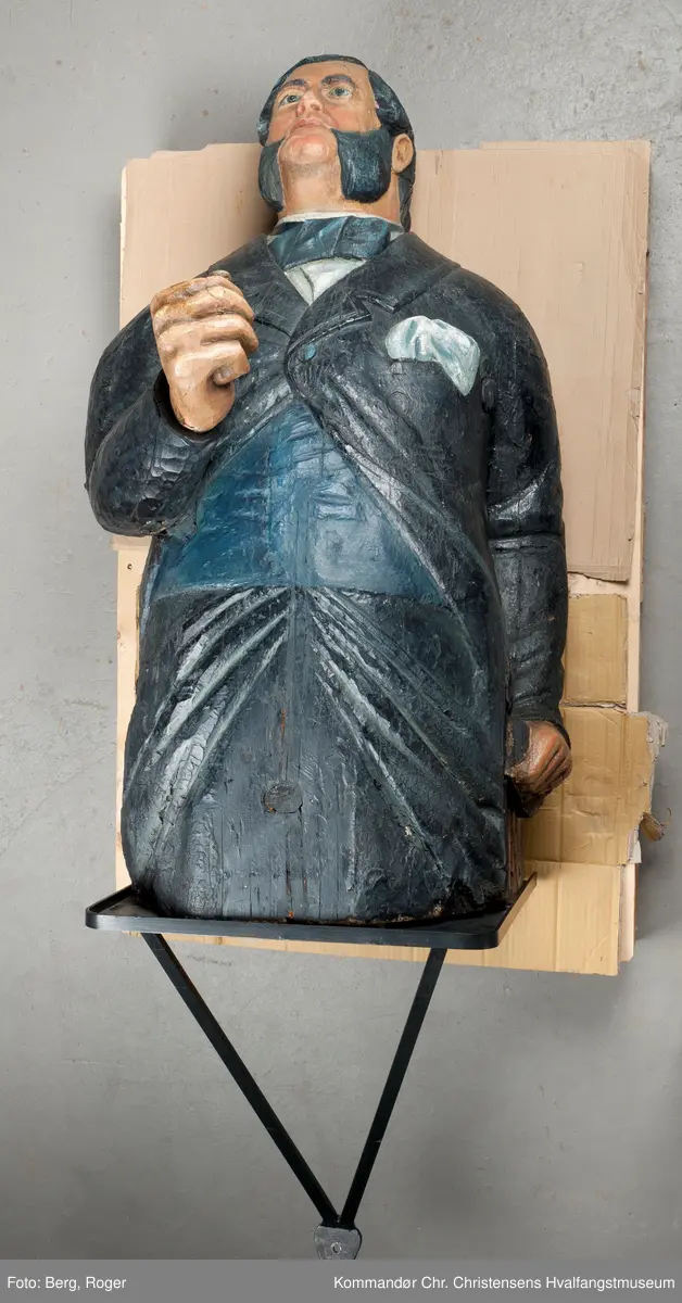 Sortkledd mann med stiv snipp. Bakkenbart, kinnskjegg uten forbindelse med hakeskjegg eller bart.