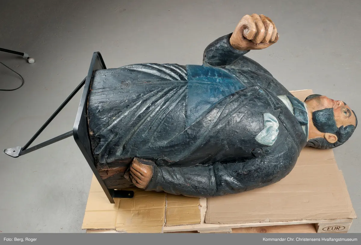 Sortkledd mann med stiv snipp. Bakkenbart, kinnskjegg uten forbindelse med hakeskjegg eller bart.