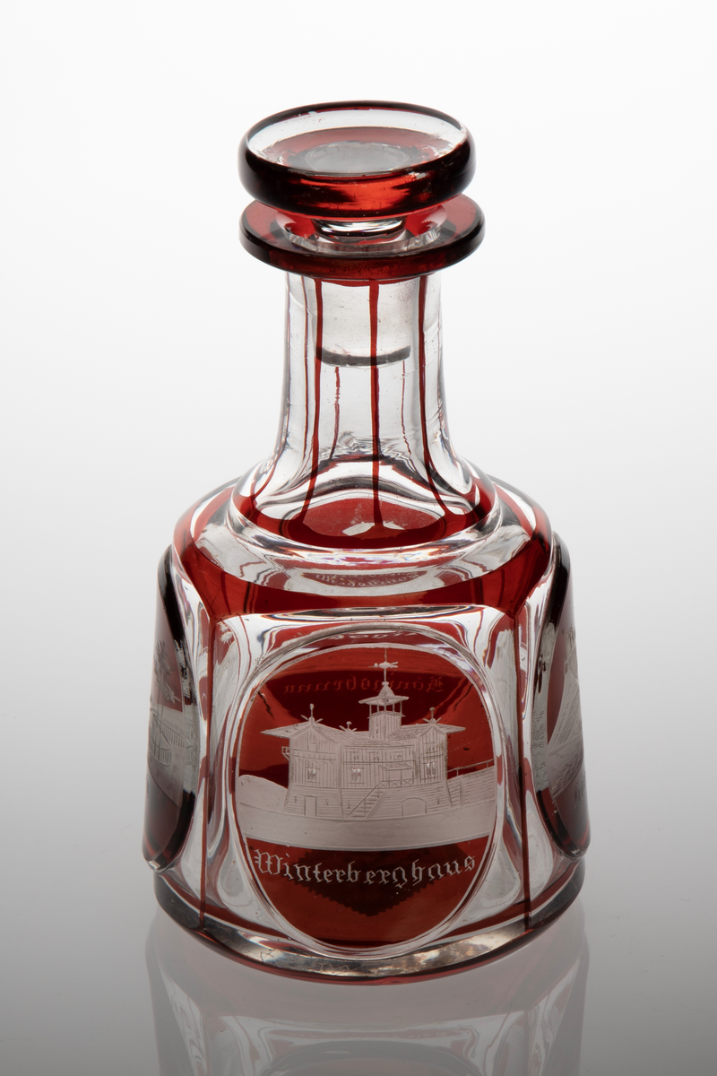 Karaffel med propp i klart glass med dekor i rødt overfang. Sylindrisk korpus med sider som skrår svakt innover. På korpus fire slipte fasetter, i hver fasett er ovalslipt medaljong, i disse gravert dekor i form av bygninger. Under respektive bygning står det: "Königsbrunn", "Basteÿ", "Winterberghaus" og "Königstein." Hals inndelt i sju seksjoner med hjelp av streker i rødt glass. Skiveformet rød propp.