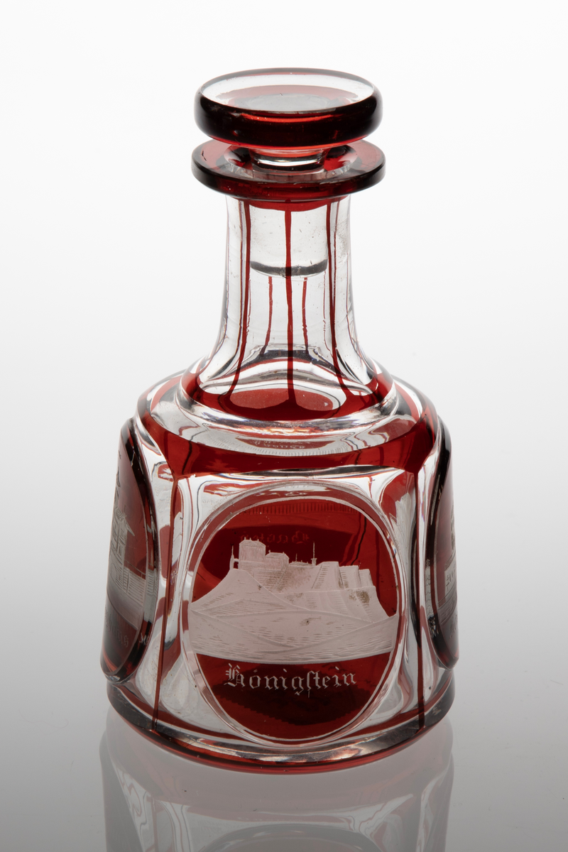 Karaffel med propp i klart glass med dekor i rødt overfang. Sylindrisk korpus med sider som skrår svakt innover. På korpus fire slipte fasetter, i hver fasett er ovalslipt medaljong, i disse gravert dekor i form av bygninger. Under respektive bygning står det: "Königsbrunn", "Basteÿ", "Winterberghaus" og "Königstein." Hals inndelt i sju seksjoner med hjelp av streker i rødt glass. Skiveformet rød propp.