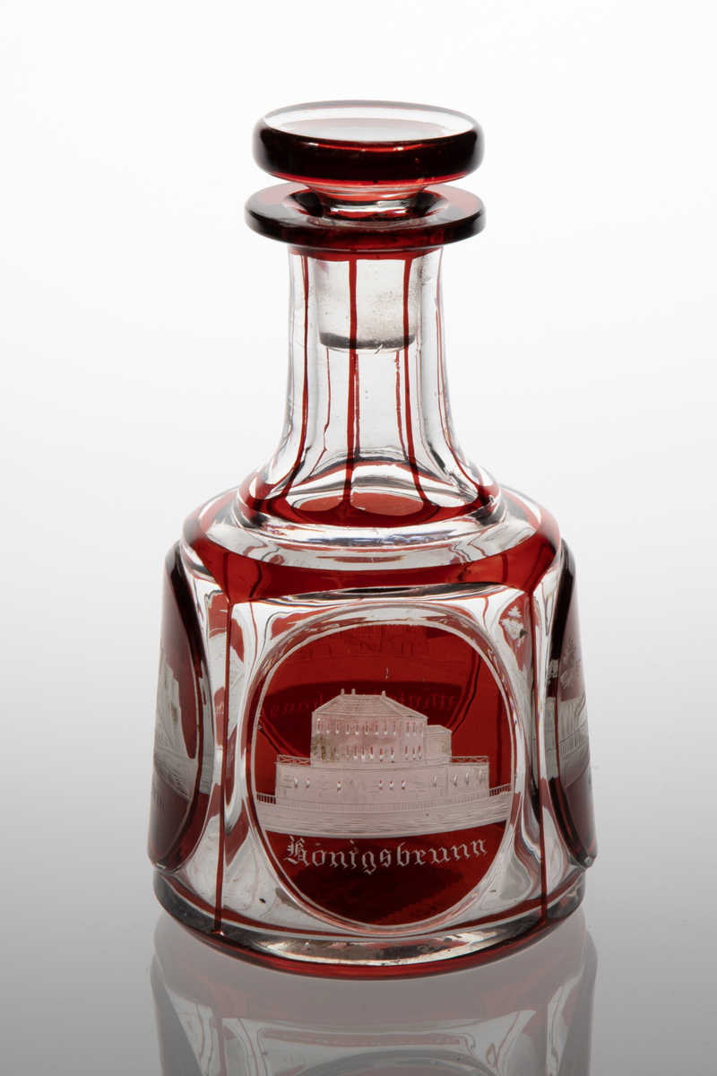 Karaffel med propp i klart glass med dekor i rødt overfang. Sylindrisk korpus med sider som skrår svakt innover. På korpus fire slipte fasetter, i hver fasett er ovalslipt medaljong, i disse gravert dekor i form av bygninger. Under respektive bygning står det: "Königsbrunn", "Basteÿ", "Winterberghaus" og "Königstein." Hals inndelt i sju seksjoner med hjelp av streker i rødt glass. Skiveformet rød propp.