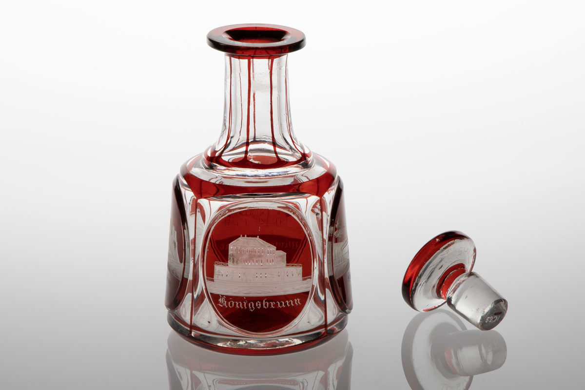 Karaffel med propp i klart glass med dekor i rødt overfang. Sylindrisk korpus med sider som skrår svakt innover. På korpus fire slipte fasetter, i hver fasett er ovalslipt medaljong, i disse gravert dekor i form av bygninger. Under respektive bygning står det: "Königsbrunn", "Basteÿ", "Winterberghaus" og "Königstein." Hals inndelt i sju seksjoner med hjelp av streker i rødt glass. Skiveformet rød propp.