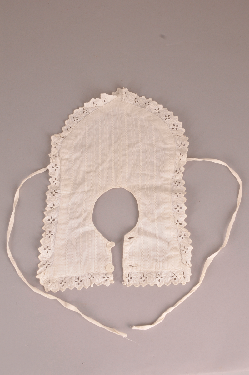 Barnesmekke i kvitt bomullstøy. Mønster på den eine sida.Blonde kring kanten. Avrunda framme. To knappar og knapphol bak.Knytband i bomull framme. Sauma på maskin.