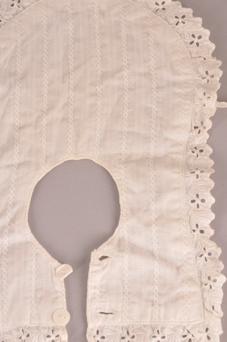 Barnesmekke i kvitt bomullstøy. Mønster på den eine sida.Blonde kring kanten. Avrunda framme. To knappar og knapphol bak.Knytband i bomull framme. Sauma på maskin.