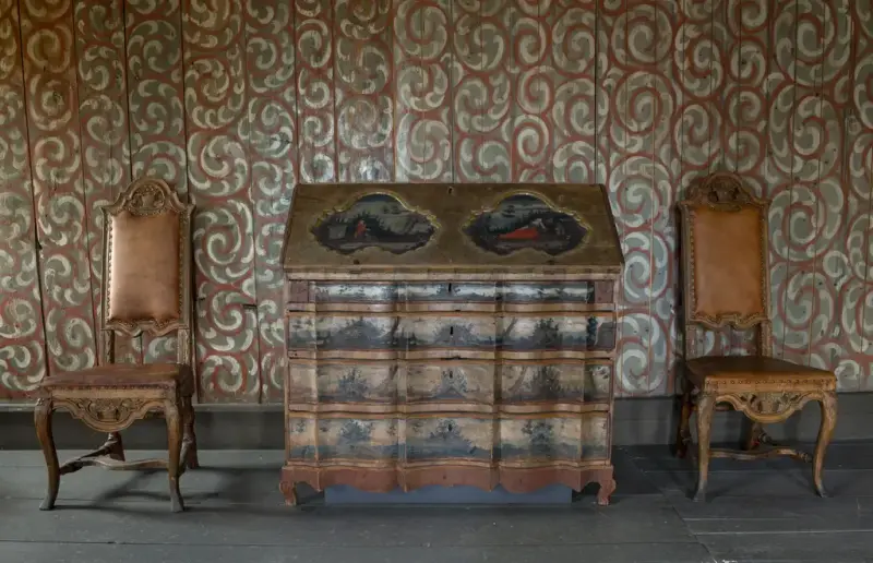 To høyryggede stoler medx sirlige utskæringer og pute og rygg i brunt skinn står på hver sin side av et dekormalt skatoll. Vegge bak har rød, grågrønn og hvit dekormaling i snirkelmønster.