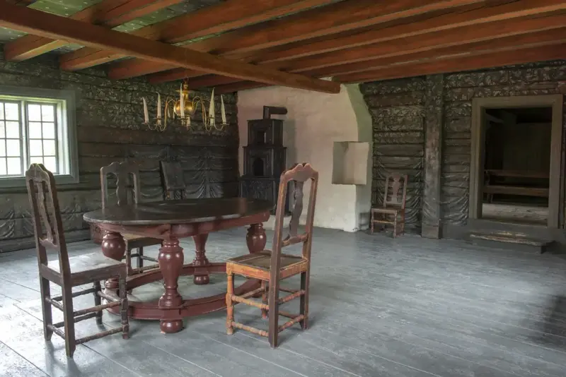 3 trestoler med høye rygger og litt slitt brunmaling står rundt et ovalt trebord med store kuler på bena, midt i et rom. Over bordet henger en messinglysekrone med lange lys ned fra tak med røædmalte dragere. Golvet er malt brlågrønt, og veggene har samme bunnfarge, men med draperimaling. Peisovn i hjørnet.