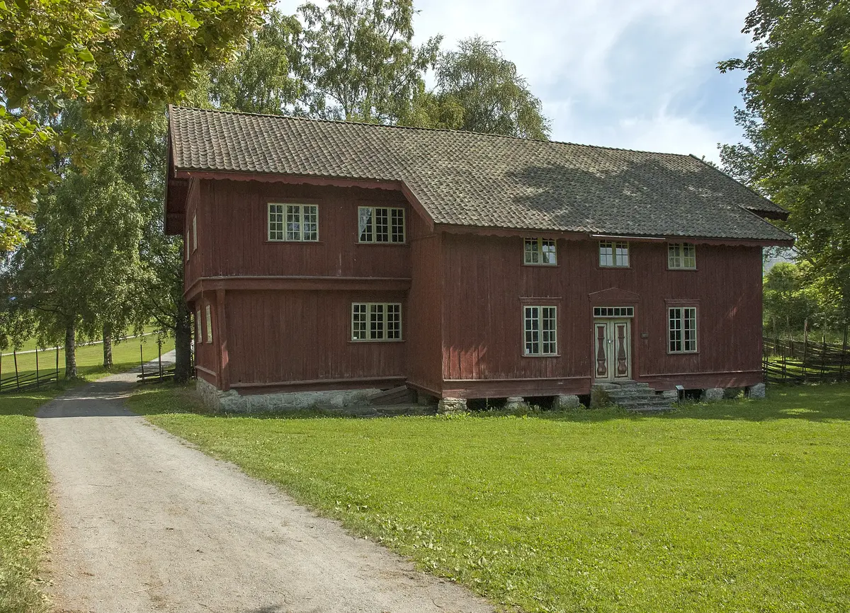 Rød, plankekledd bygning fra 160, med litt utstikkende andreetasje og innbygget svalgang som stikker ut. Smårutete vinduer og enkrums terrakottabrun takstein.