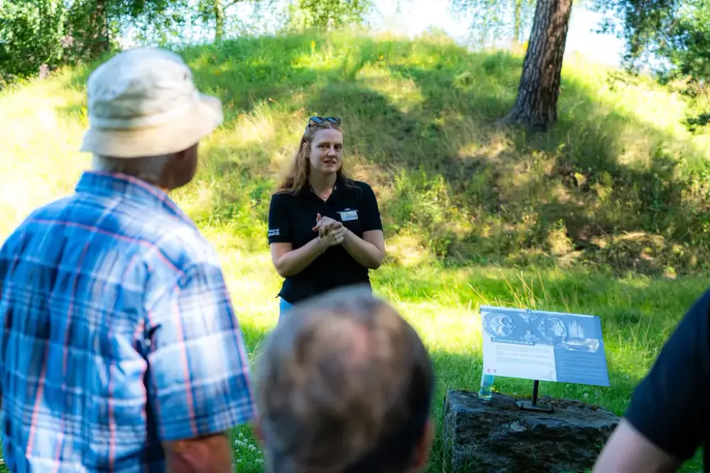 Engasjert guide står foran sommer-grønn gravhaug med tilhørere.