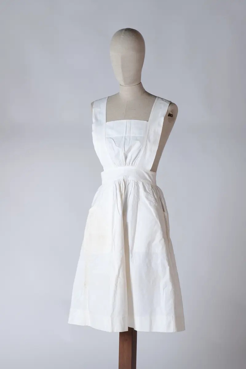 Del av sykepleieruniform bestående av to deler, kjole og forkle. Forkleet er av hvit bomull med to store lommer og det dekker kjolen helt ned til falden. Det har knappelukking bak på ryggen med 3 store knapper. Stroppene til forkleet går i kryss på ryggen.