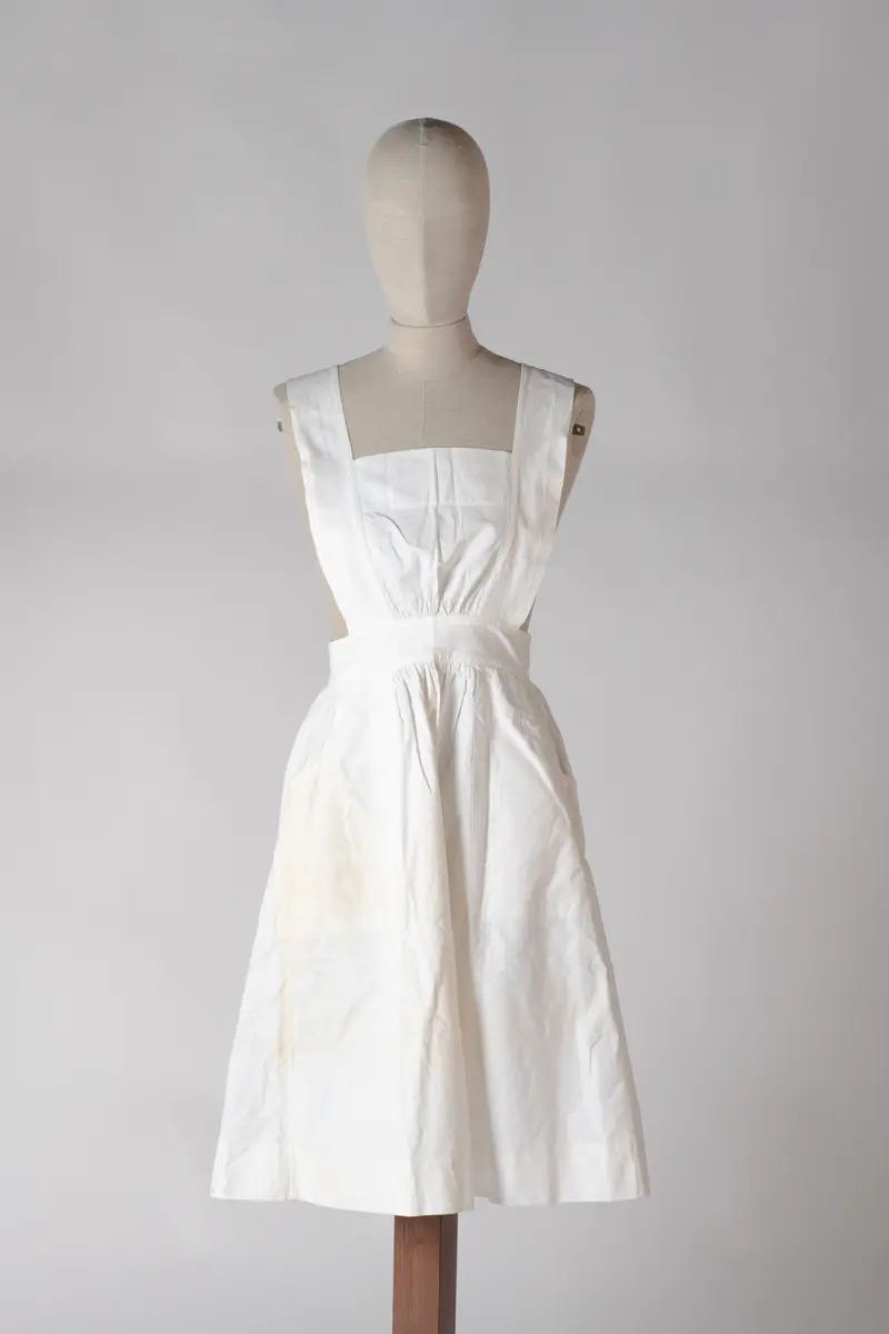 Del av sykepleieruniform bestående av to deler, kjole og forkle. Forkleet er av hvit bomull med to store lommer og det dekker kjolen helt ned til falden. Det har knappelukking bak på ryggen med 3 store knapper. Stroppene til forkleet går i kryss på ryggen.