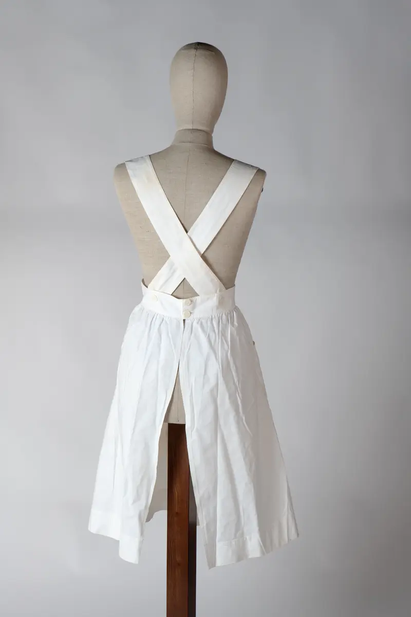 Del av sykepleieruniform bestående av to deler, kjole og forkle. Forkleet er av hvit bomull med to store lommer og det dekker kjolen helt ned til falden. Det har knappelukking bak på ryggen med 3 store knapper. Stroppene til forkleet går i kryss på ryggen.