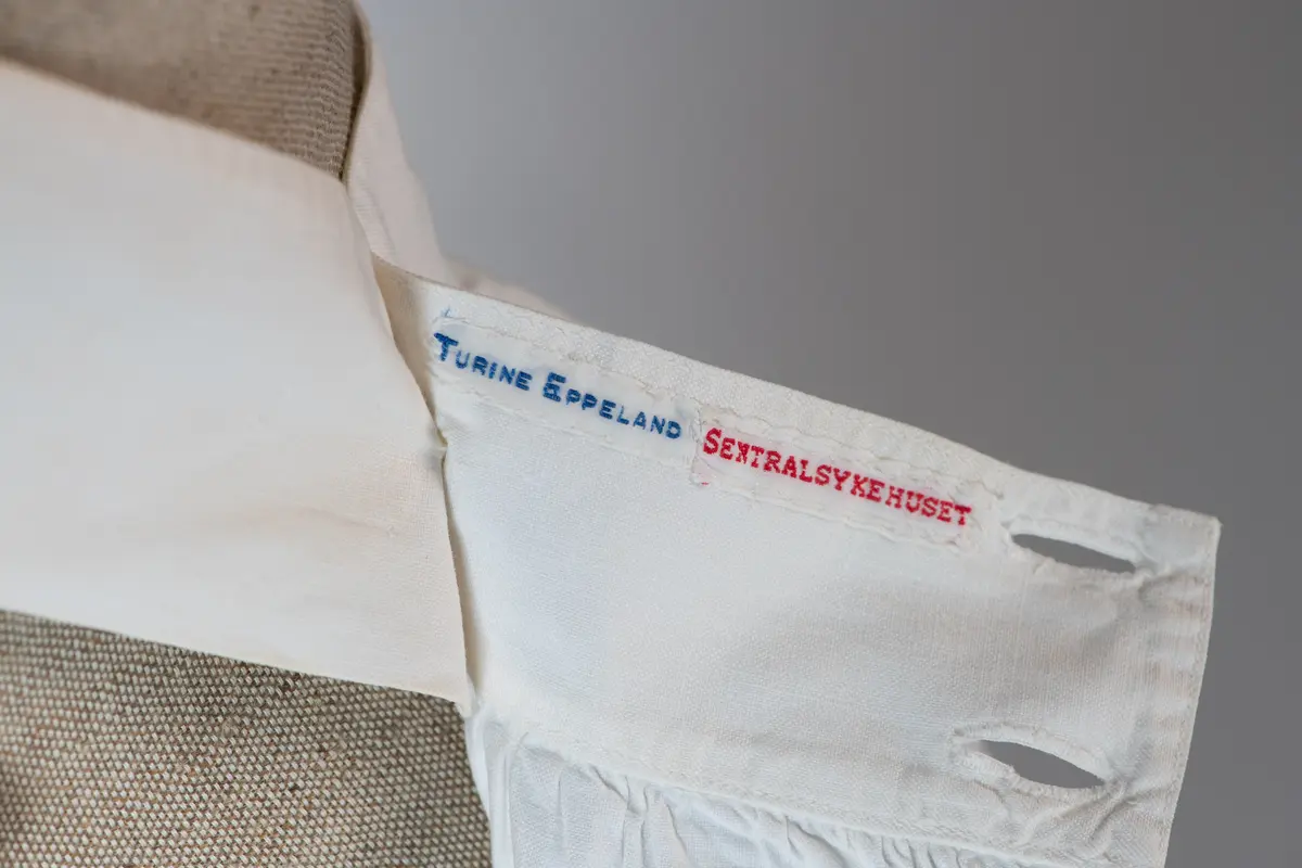 Del av sykepleieruniform bestående av to deler, kjole og forkle. Forkleet er av hvit bomull med to store lommer og det dekker kjolen helt ned til falden. Det har knappelukking bak på ryggen med 3 store knapper. Stroppene til forkleet går i kryss på ryggen.