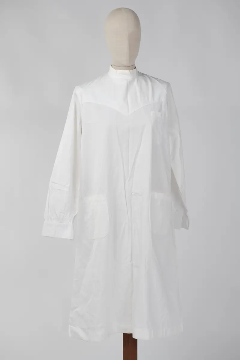Lang sykepleieruniform i hvit bomull. Uniformen har høy hals med 4 knapper som går halvveis ned på ryggen, og manskjett på ermene med 1 knapp. På forsiden er det 1 liten lomme øverst på venstre side og 2 større lommer på midten av uniformen.