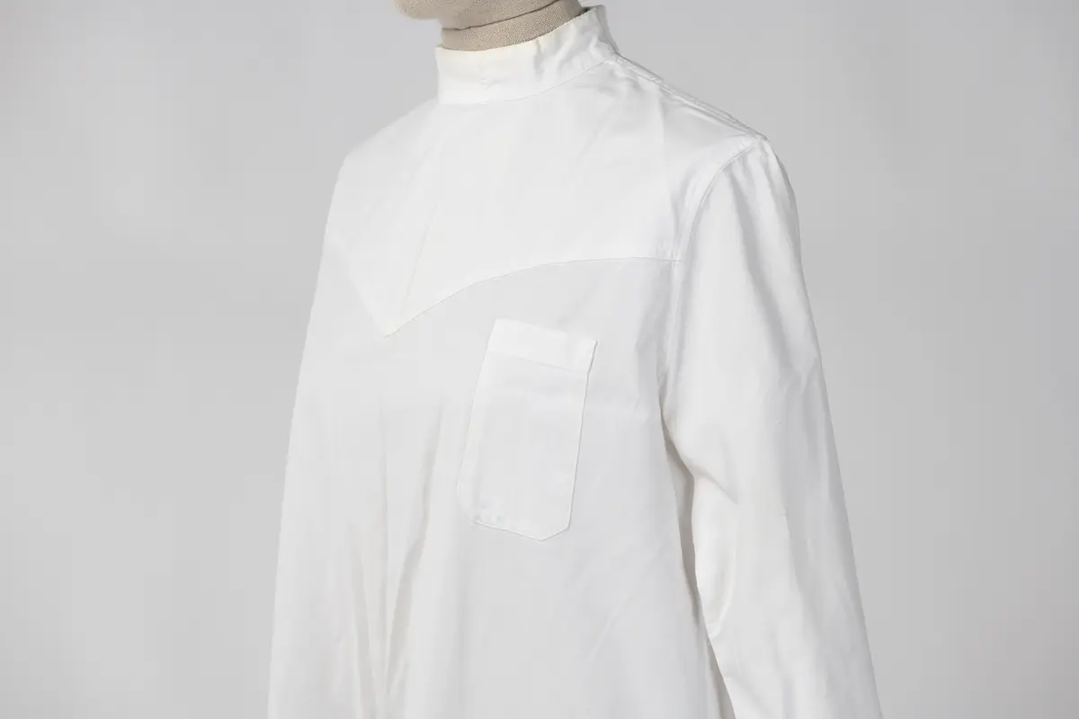 Lang sykepleieruniform i hvit bomull. Uniformen har høy hals med 4 knapper som går halvveis ned på ryggen, og manskjett på ermene med 1 knapp. På forsiden er det 1 liten lomme øverst på venstre side og 2 større lommer på midten av uniformen.